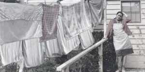 A woman in depression era clothing stands outside a house with her hand to her head, a full clothesline of washing to her left