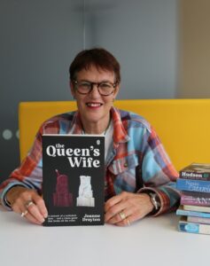 Larger image link: Joanne Drayton, pictured next to a pile of her book picks and holding her own book, The Queen's Wife