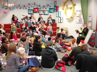A librarian leads a Baby Rock and Rhyme session in a brightly-decorated library. A large crowd of parents and babies are in attendance, some sitting on the ground, others on the terraces.
