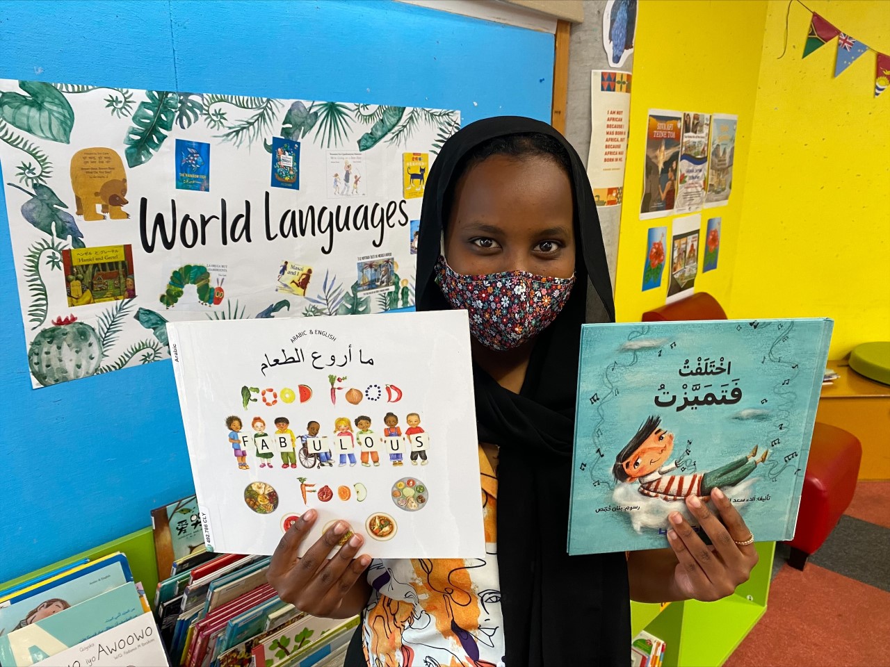 A smiling librarian, wearing a colourful facemask and hijab, is holding two beautiful picture books in Arabic. Behind her, you can see the children's world languages collection at Newtown Library, with lots of books in colourful shelving and comfortable seats nearby.