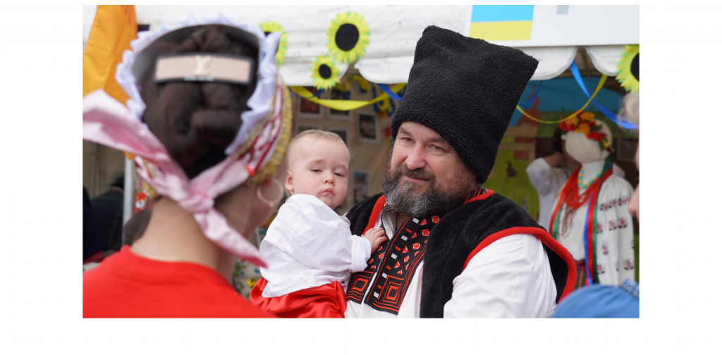 A man in Ukrainian cultural dress, including a tall fur cap and an elaborately-knotted brocade, is holding a small child in front of a festival stall which is decorated with sunflowers.