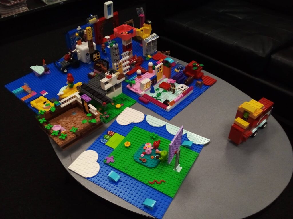 Several colourful LEGO constructions sitting on a table. Among them are several blue-and-green landscapes, a house, a fire engine, and a garden. 
