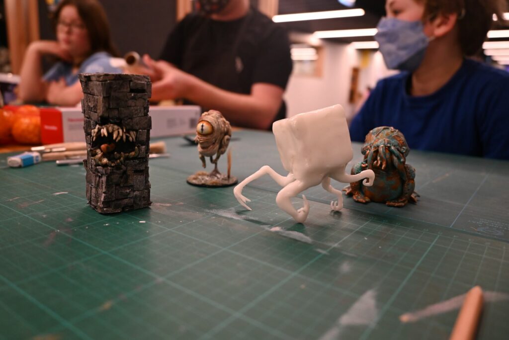 Four small sculpted figurines sit on a green cutting mat. From left to right, they appear to be a sentient brick wall, a one-legged, one-eyed monster, a white block-like creature with tentacle-like arms, and a frog-like creature with tentacles coming out of its mouth