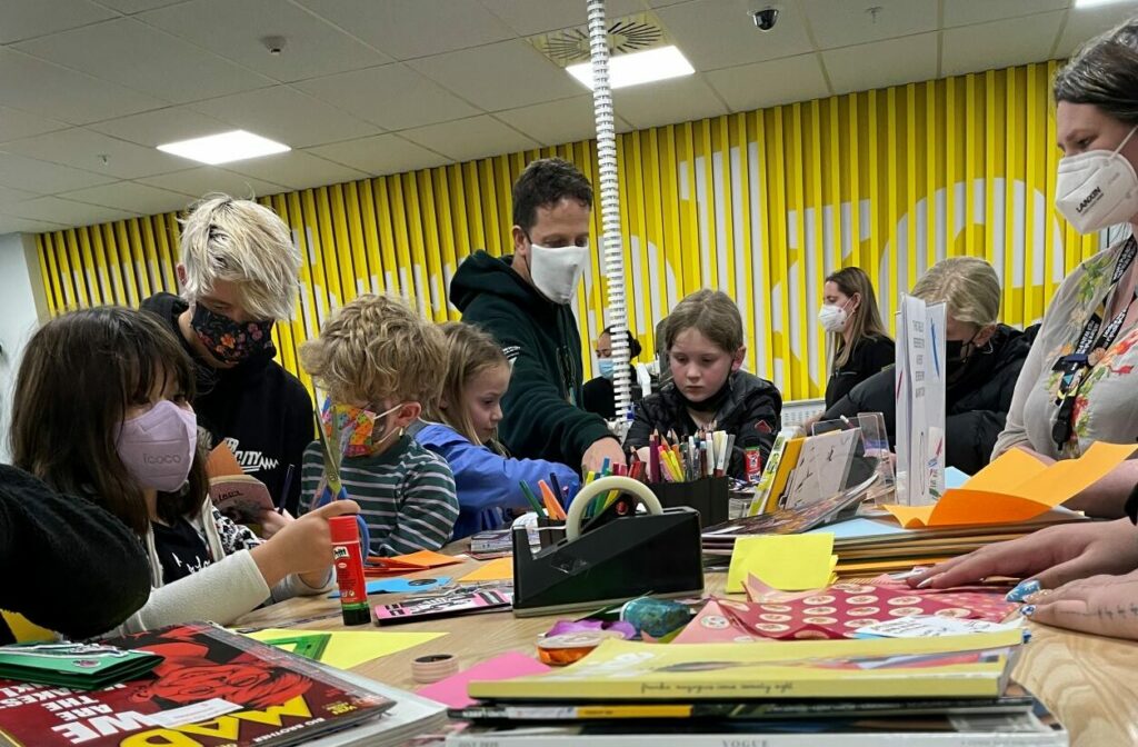 A large group of children gathered around a table.  On the table are various craft supplies, magazines, pens and pencils.