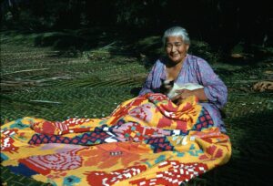 A photo of a woman sitting on a woven harakeke mat, weaving a colourful mat.