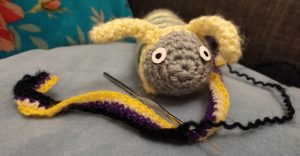 Claude, a grey, green, and yellow caterpillar is sitting on a cushion crocheting the last row of a scarf. The stripes of the scarf are, in order, yellow, white, purple, and the last one is black.