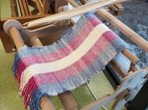 A newly finished scarf lies folded on the small loom in the Johnsonville makerspace. The stripes on the scarf go (from left to right) blue, pink, white, pink, blue.