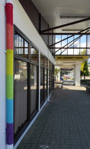 A picture taken looking down the street towards the Tawa Community Centre entrance. It is a sunny day. Lining up with the left side of the picture is a pipe attached to the building, that has is wearing a rainbow cover.