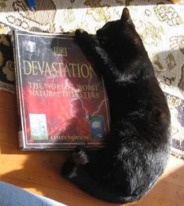 A black cat is lying curled around the right side of a large book. his teeth and front claws are wrapped around the top corner. The title of the book is Devastation: the world's worst natural disasters
