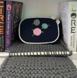 Image shows a floral embroidery set in black fabric, resting on a piece of gray knitting, sitting on a bookshelf between several books.