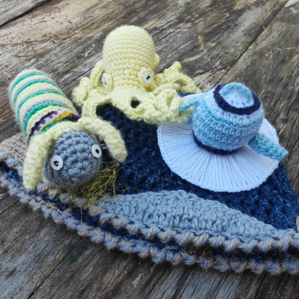 A green, grey, and yellow crocheted caterpillar sits next to a yellow crocheted octopus. The octopus has one tentacle through the handle of a white and blue crocheted teapot.
