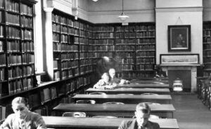 Black and white photograph, lines of tables and bookcases lining the walls. Someone at a table is lit up by light coming through a window, behind them are two blurry figures