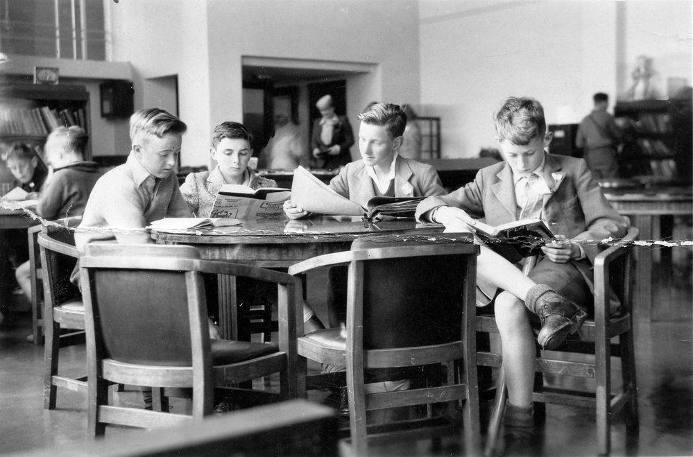 Four boys in their early teens sit around a table reading newspapers and large books
