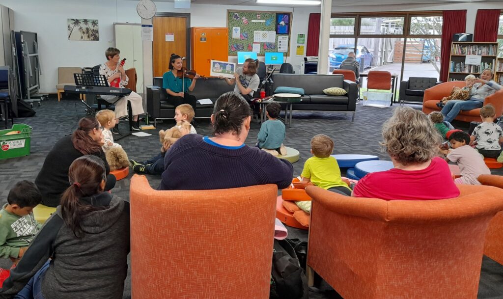 Group of children and parents watch librarians reading book, with musical instruments