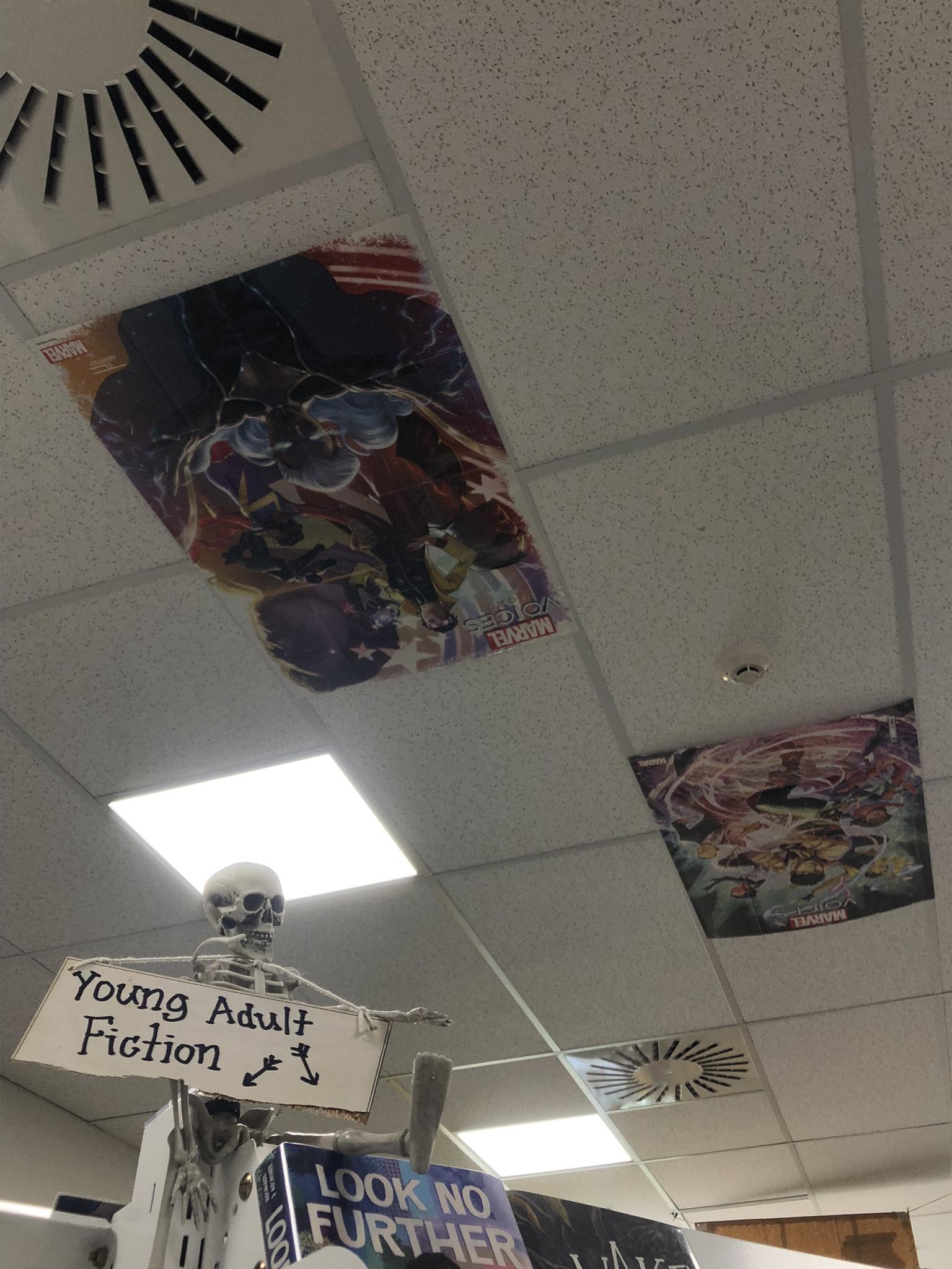 A plastic skeleton perches on a bookshelf with a sign reading 'Young Adult Fiction'. On the ceiling tiles above are movie posters.