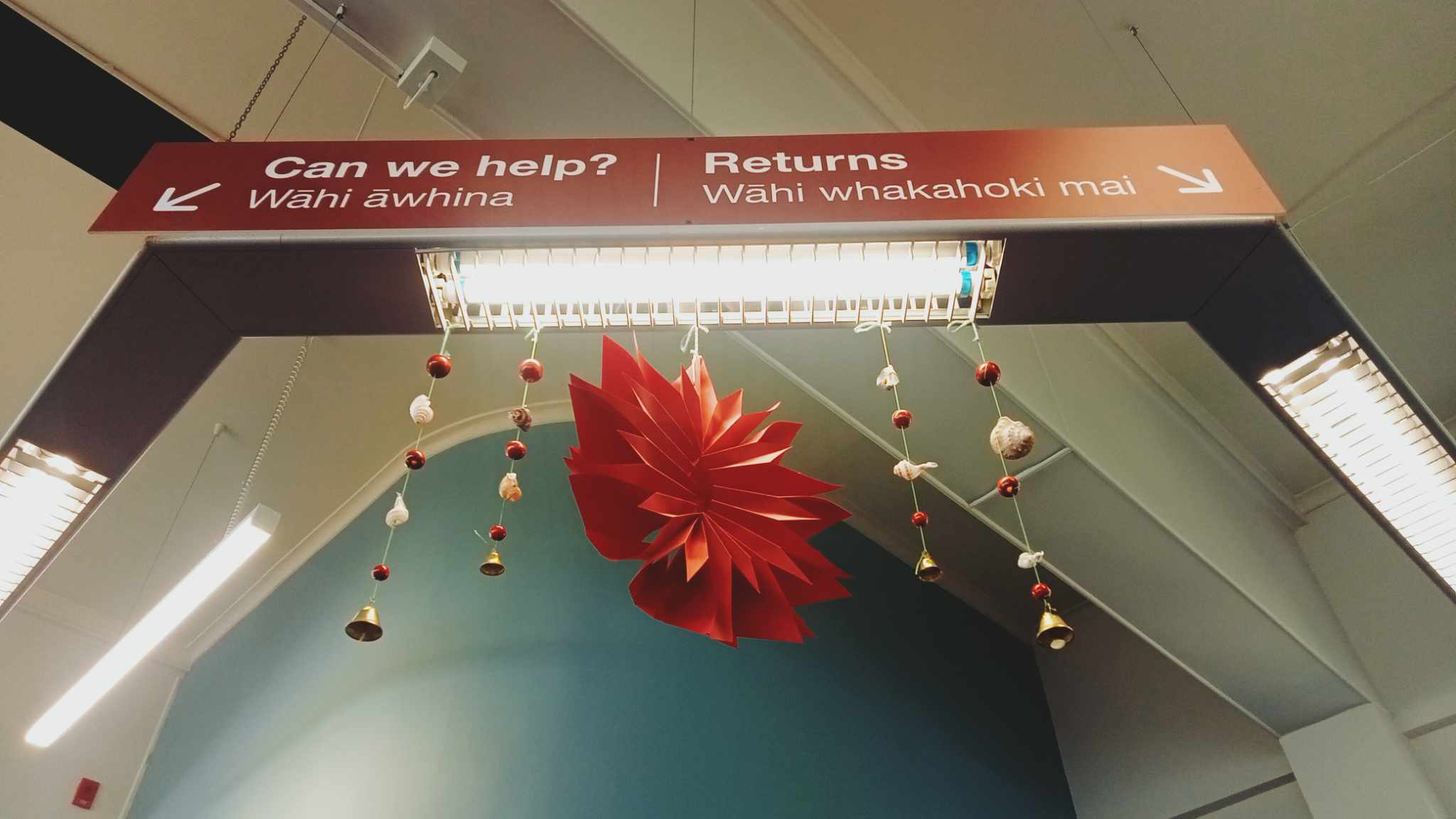 A large red paper flower and strings of red beads and white shells hanging from a red library sign with 'Can we help" written on it.