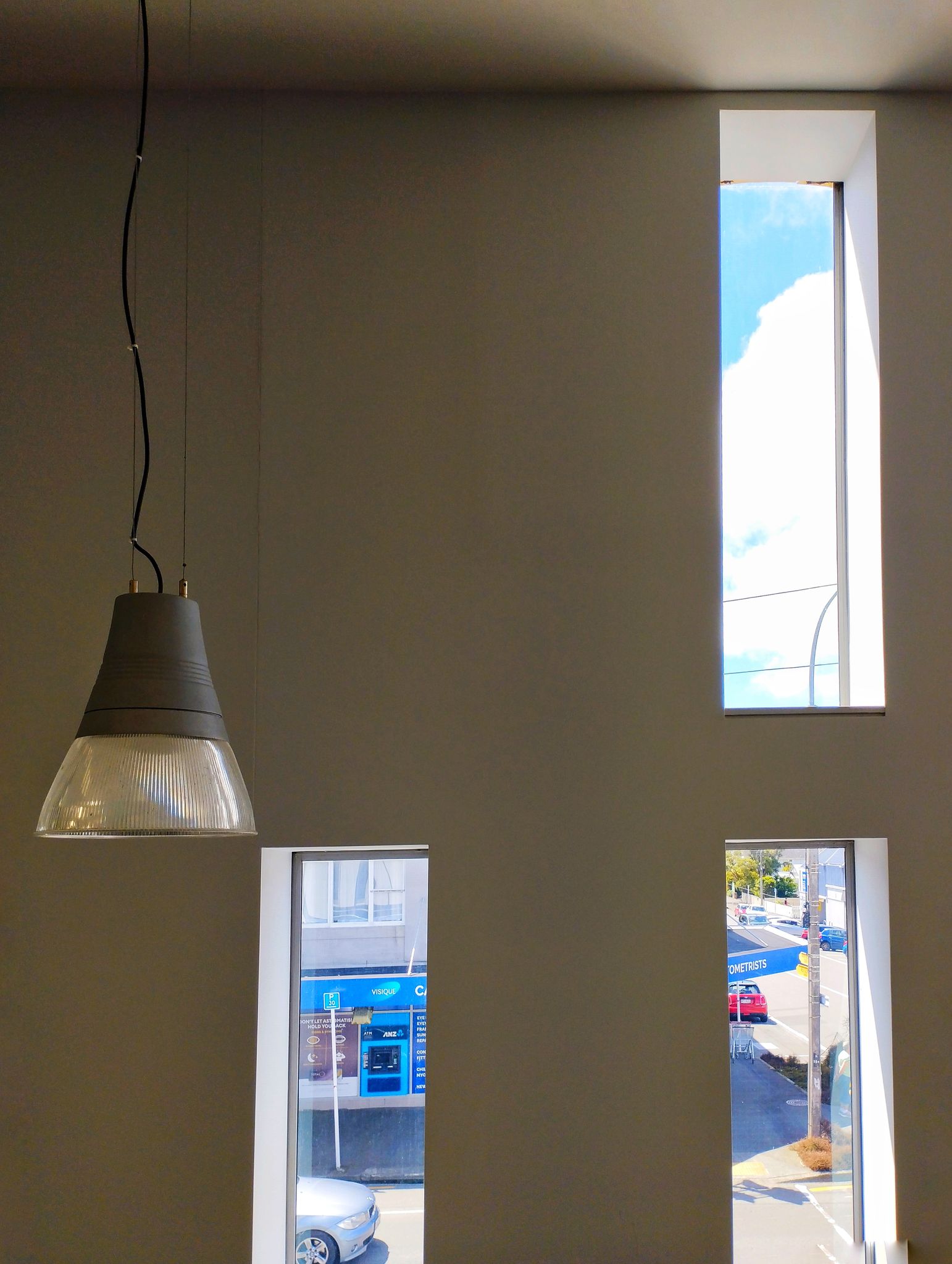 Three narrow windows in a white wall look out onto the street and blue sky. A conical light hangs on the left, the bottom level with the top corner of a narrow window.