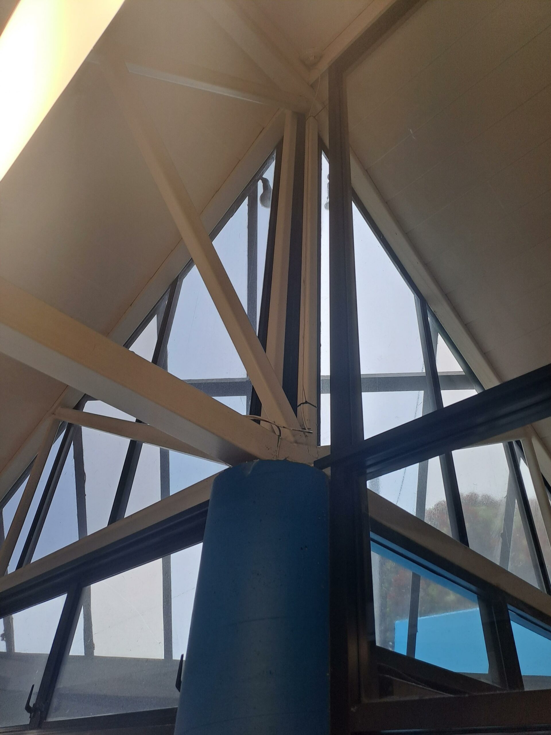 A blue pillar with outreaching metal struts atop it, in front of a corner window.