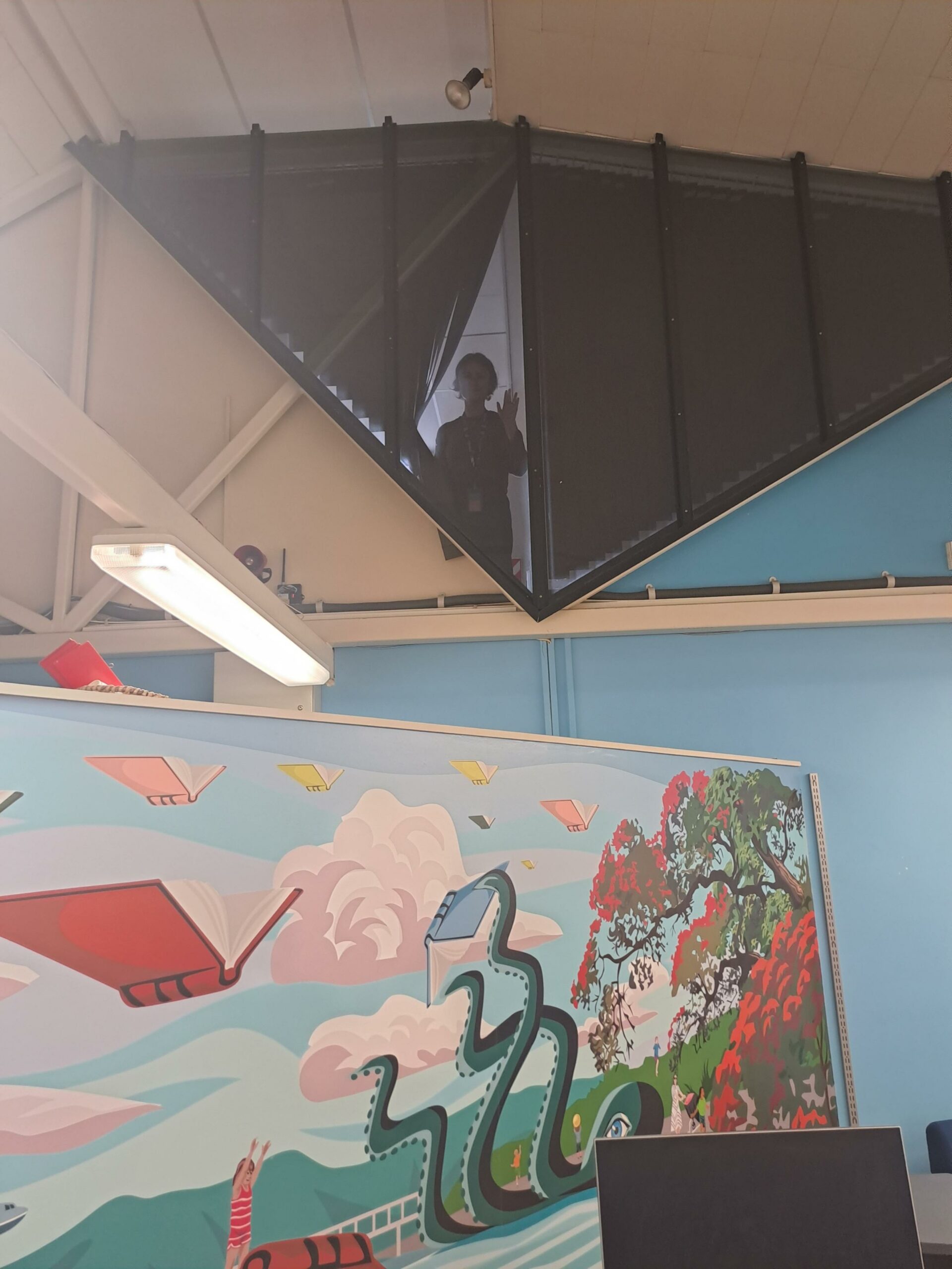 A triangular internal window above a mural of a beach with swimming child, reading octopus, and books flying through the air. In the window, a silhouette of a librarian waves.