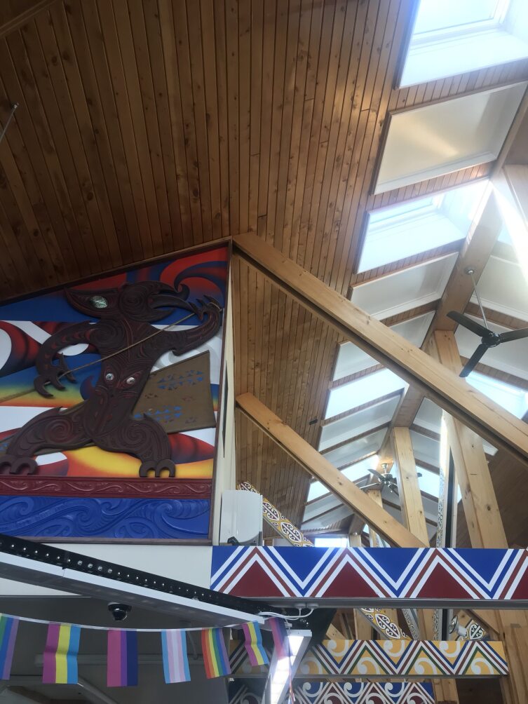 Wooden beams on the right angle up to the natural wooden ceiling. On the left a wooden taniwha attached to a colourful painted wall looks up towards the peak of the ceiling.