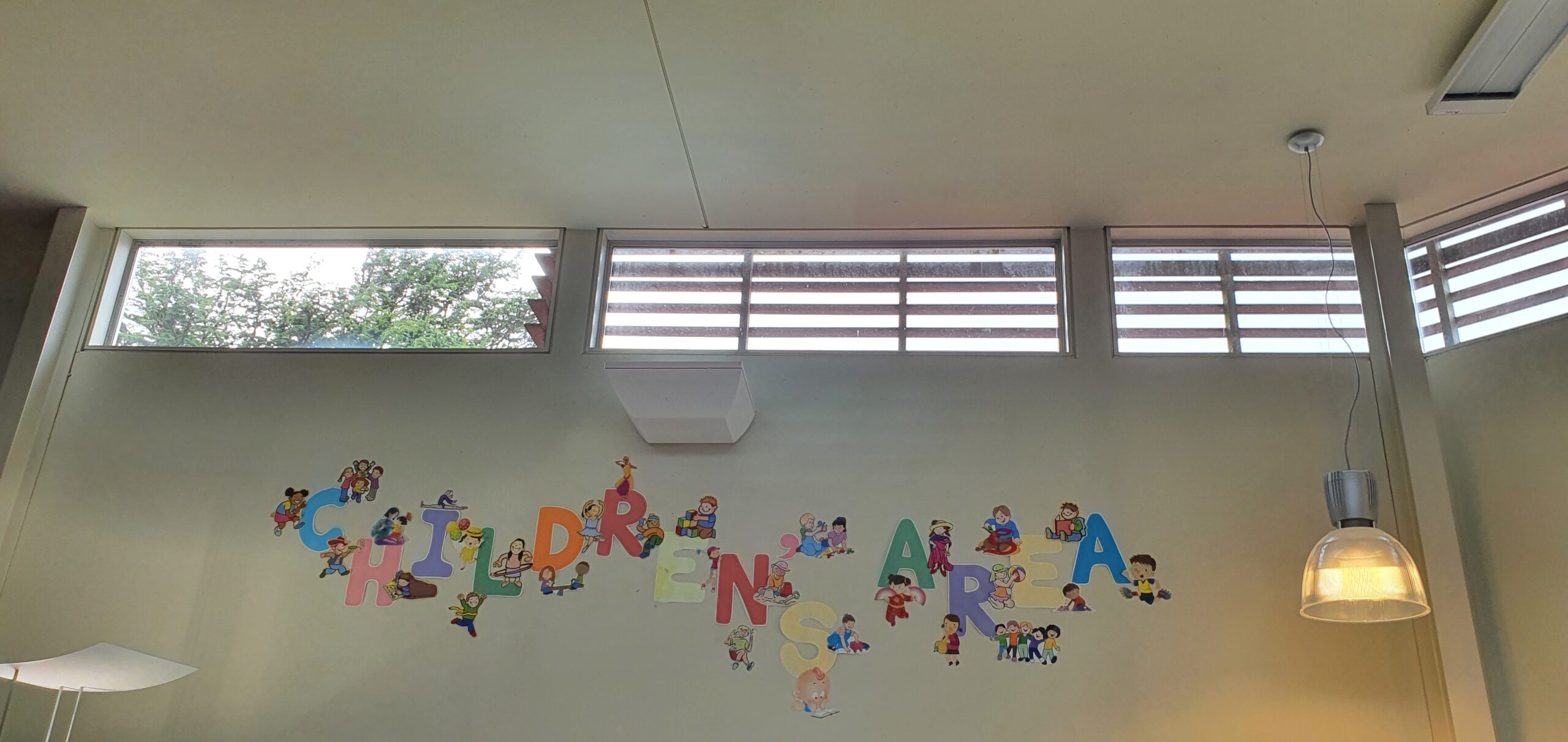 Coloured paper letters spelling 'Children's Area' on the wall below small high windows with wooden blinds.