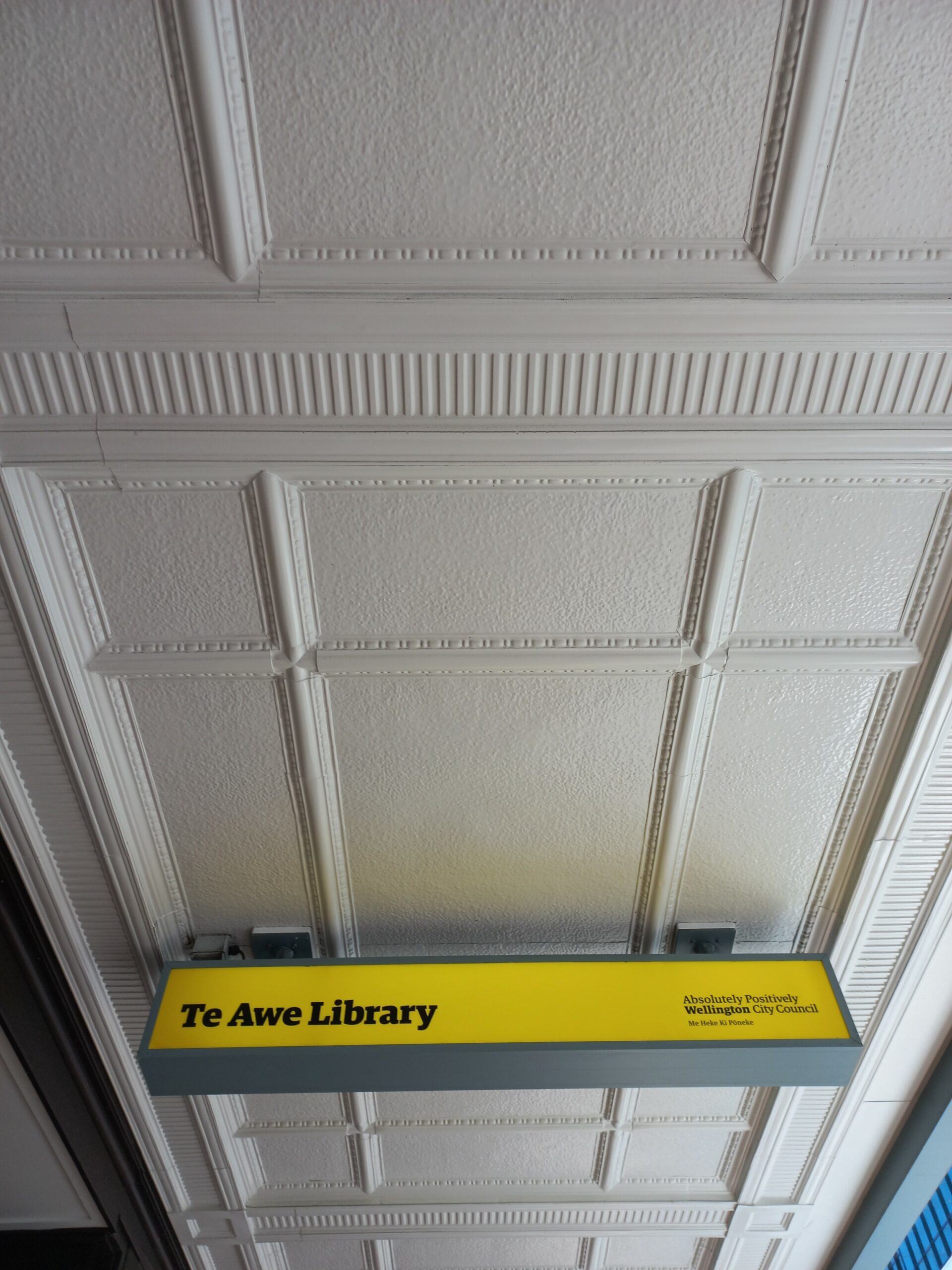 Decorated 1900s eave overhanging the footpath, with a yellow 'Te Awe Library' sign hanging from it.