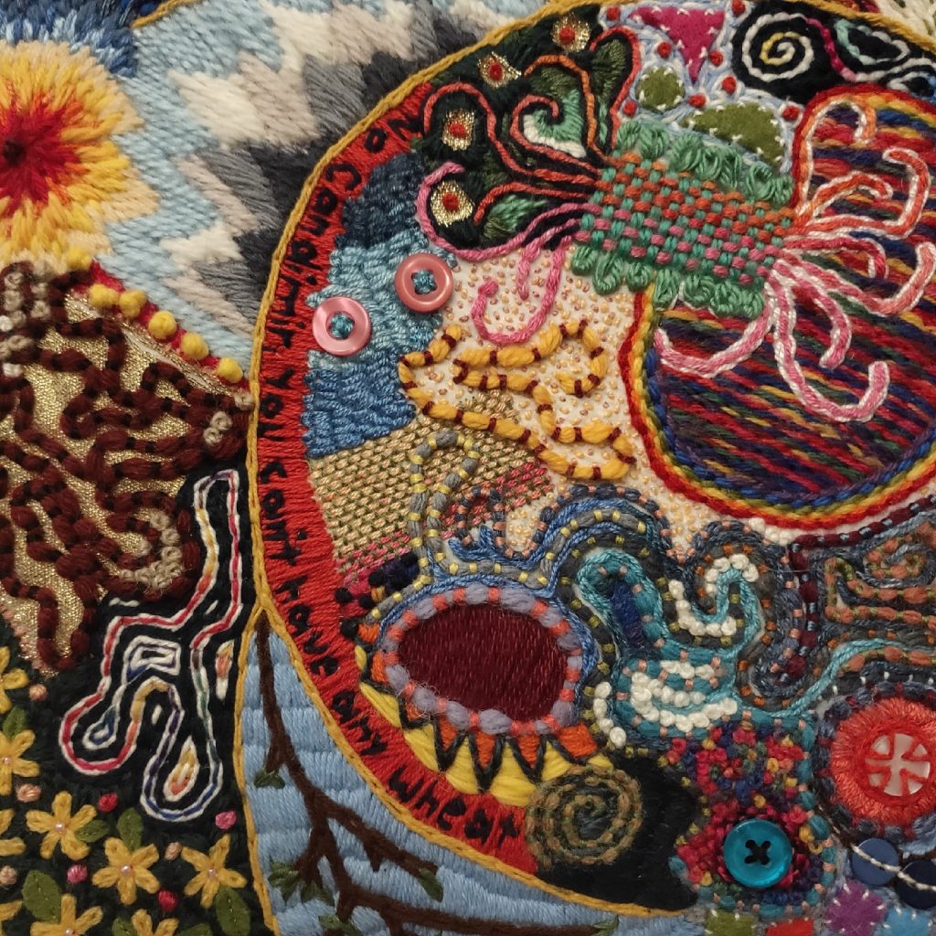 A chaotic piece of embroidery. Black letters on a red background across the centre read "No Candimir, you can't have any wheat". There are mountains in the upper left corner, and yellow flowers on a dark green background in the lower left. Some beads and buttons are sewn in on the right side, and the whole photo area is covered in colourful stitches.